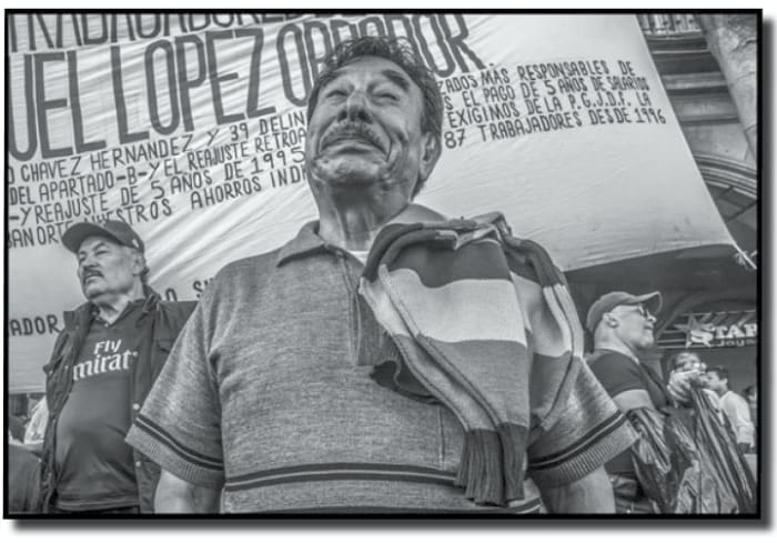 Among the Mexicans in the Zocalo, celebrating the inauguration of Mexico's new President, Andres Manuel Lopez Obrador, are workers fired in the privatization of the city's Ruta 100 busline over twenty years earlier, at the beginning of the NAFTA era. Many workers like these believed AMLO's promise that he would end the neoliberal policies of the NAFTA era. (David Bacon, Stanford University Green Library)