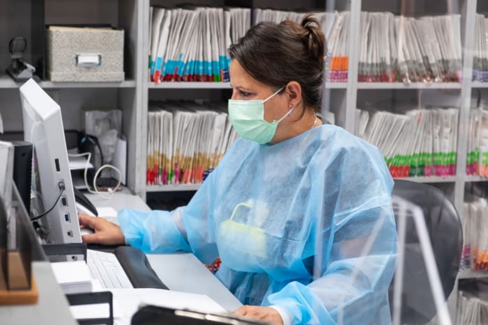 Hispanic receptionist at dental office wearing mask and gown for protection during the Covid-19 pandemic. She is working at the computer with patient files in the background.