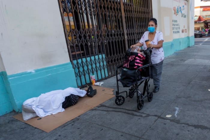 LOS ANGELES, CA - MAY 08: A woman carrying a load of bread walks near a homeless person sleeping on the sidewalk as businesses in the flower district in Skid Row reopen in time for Mother's Day on May 8, 2020 in Los Angeles, California. Local stay-at-home restrictions are being relaxed to allow the reopening of some businesses, including bookshops, clothing stores, car dealerships, and some low-risk retailers that can provide curbside pickup. Many recreational areas like parks, trails and golf courses are set to reopen tomorrow as well. However, L.A. mayor Eric Garcetti has repeatedly warned that if people do not wear masks or face coverings and practice social distancing at these businesses and locales, the city may have to close them down once again. (Photo by David McNew/Getty Images)