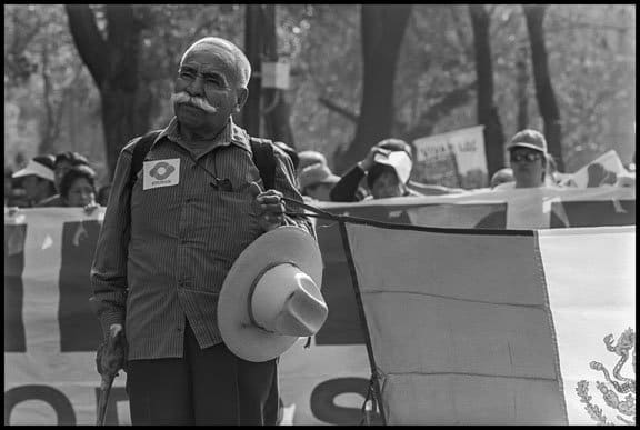 An older worker holds the Mexican flag, and wears the insignia of the university workers union STUNAM.