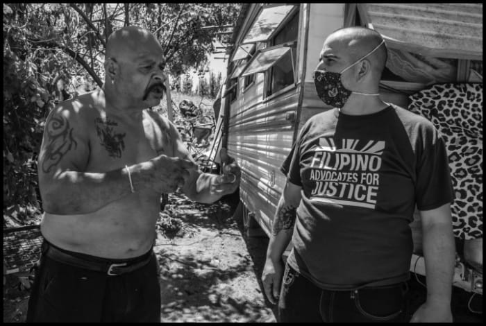 POPLAR, CA - 12JULY20 - Rosendo and Josefina live in a trailer in an encampment on the Tule River levee near Porterville. Rosendo was released from prison just eight months earlier, after being incarcerated for 38 years. The moved the trailer to the levee because they have no money to pay rent. Rosendo talks with Arturo Rodriguez, an organizer with the Larry Itliong Resource Center, about conditions in the camps along the river. Copyright David Bacon