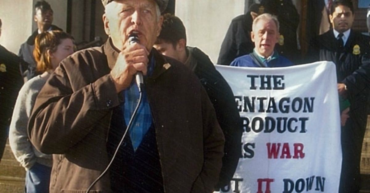 Phil Berrigan in front of a "wreckage of war" die-in at the River Entrance. Frida is half-visible behind to the left. Photo taken by Rick Reinhard on December 30, 1996. The banner reads, "The Pentagon Product Is War: Shut It Down." (Photo via Frida Berrigan)