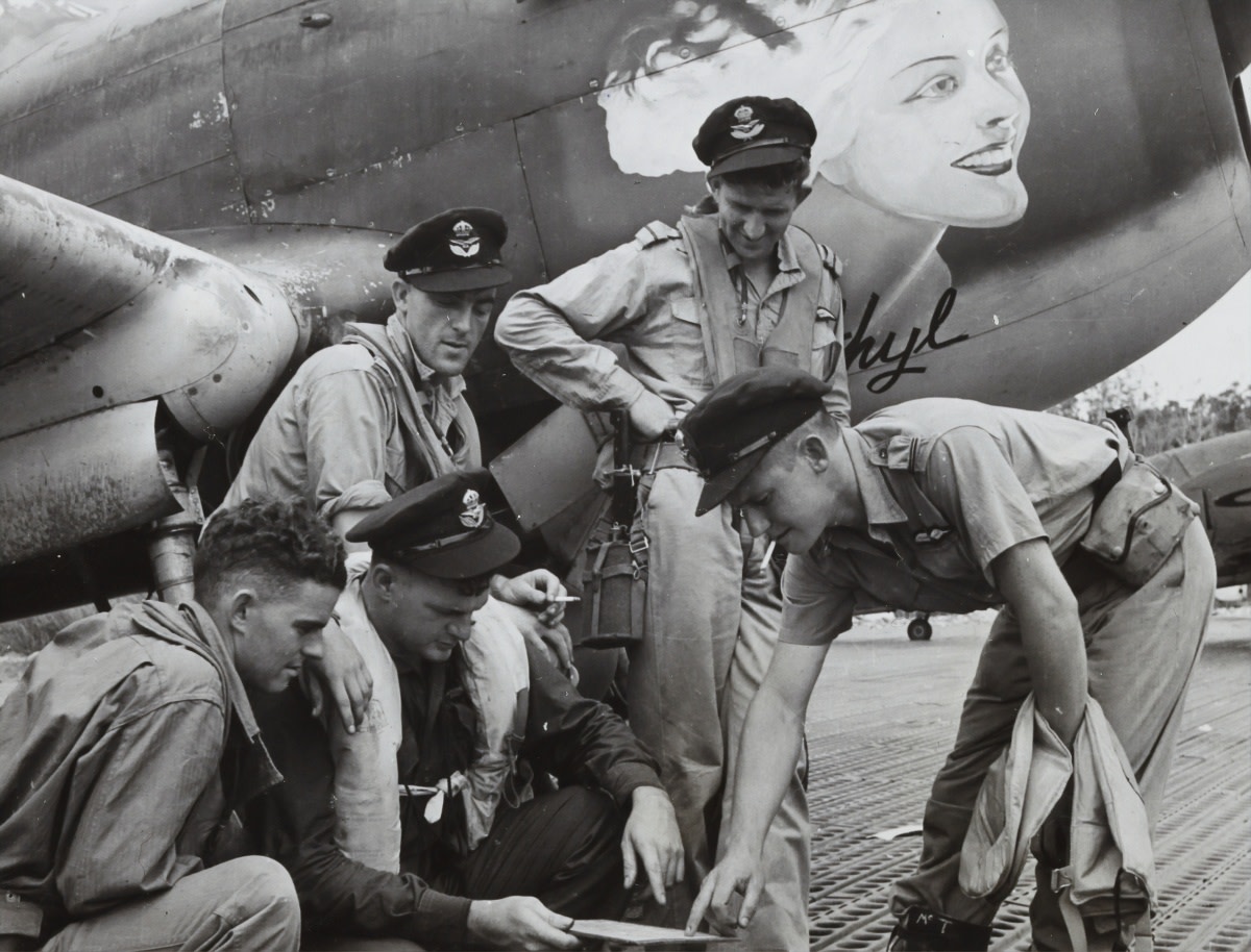 1941. A group of 77 Squadron pilots posed next to a P-40 Kittyhawk aircraft looking at a board. The nose art of a blonde woman can be seen in the background. Goodenough Island, New Guinea (Photo by Museums Victoria on Unsplash)