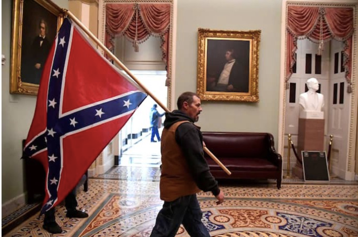 During the January 6th insurrection, a photographer captured this image of a rioter carrying the Confederate battle flag inside the Capitol. The oil portrait on the wall is of Senator Charles Sumner, a Massachusetts abolitionist who was nearly beaten to death in 1856 for his anti-slavery views.         Credit: Mike Theiler/Reuters.